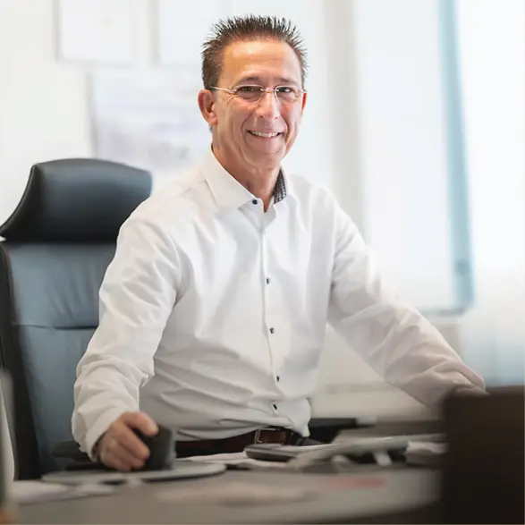 Mann sitzt an einem Schreibtisch mit Computer und Unterlagen in einem Büro und blickt zur Kamera